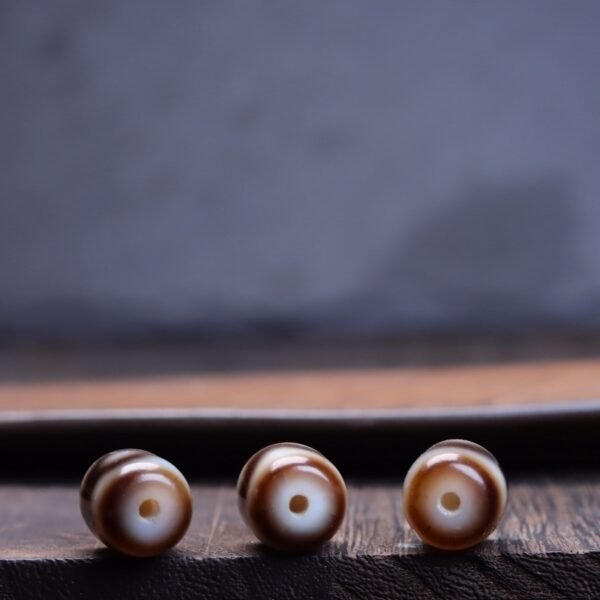 Natural Tibetan dzi bead, tiger tooth three-eye dzi, barrel-shaped spacer bead, yellowish-brown dzi bead, jewelry making supplies, Buddhist prayer beads, authentic weathering marks, Tibetan cultural artifact, spiritual symbol bead, handmade dzi bead, vintage-inspired jewelry, earthy tone spacer, mystical patterns, top bead for necklaces, bracelet accent bead, cultural craft material, protection symbol bead, wisdom dzi design, artisan jewelry component, collectible spiritual bead.